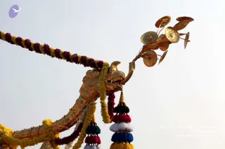 A close view of a gilded ceremonial structure. The head bears a fan-shaped cluster of gold discs and ornaments extending upward against a pale sky. Garlands of magenta and yellow-orange flowers drape across the top, and hanging tassels in green, magenta, white, blue and yellow dangle below. No people are visible in the frame.