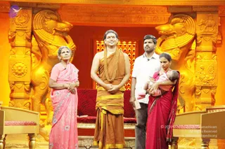 A photograph on an ornate golden stage set with carved golden lion-like pillars and a red-cushioned seat behind. A man in a saffron-brown dhoti and shawl with a long gold-toned beaded necklace stands centre, hands folded in front of him. To his left an older woman in a pink sari with blue flower print stands with her arms folded. To his right a man in a white shirt and dark trousers stands holding a paper, and beside him a woman in a red sari with a dark border holds a baby in pink clothing. Golden chairs flank the stage edges.