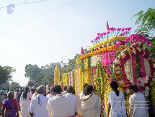 Outdoor procession on a village road under a pale sky. A large vehicle or float is completely wrapped in garlands of marigolds, white jasmine-like flowers, pink and magenta blossoms and green leaves, with small saffron and red pennants on top. Beside it, a crowd walks in the street: men in white dhotis and shirts, women in white saris and a woman in a purple sari with a red blouse at left, one person in a blue-patterned white garment at right. Overhead power lines and trees line the roadside. The scene reads as a funeral or memorial procession with a flower-decorated carriage.