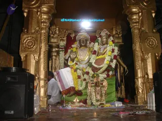 A photograph of a golden-toned pair of murti statues in a temple shrine, garlanded with white jasmine and yellow and red flower garlands. The taller figure stands with raised right hand in abhaya mudra, wearing a golden necklace and a tall crown; the second figure beside it wears a red-brown and gold garment and an ornate crown. Golden pillars frame the shrine, a green cloth is draped behind the base, and a white-clad man with a folded cloth sits at the left edge near a pot of offerings. A sign above reads Sri Anandeshwarar Samedha Anandeshwari. No living person is shown in the foreground except the seated man at left.