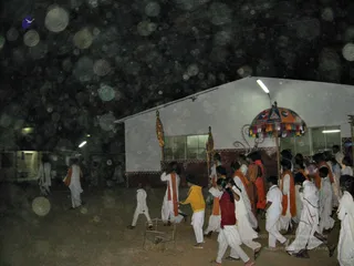 A night procession moves along a paved courtyard beside a white-walled building with fluorescent lights. Many people in white dhotis and white kurtas, several wearing orange shawls over their shoulders, walk in a line. Two long standards with gold finials are carried upright, and a large ornate umbrella with colourful tassels and a painted pattern is held aloft at right. A small metal frame cart with wheels sits in the foreground. Bright bokeh of light dots fills the dark sky above the building. Some participants wear red and yellow tops among the white clothing.