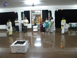 Photograph of an indoor hall with a polished reddish-brown floor at night. In the foreground a square fire pit is built of white brick blocks, with a small flame visible at its base. Behind it, a group of about nine people stand in a semicircle facing the pit: several men in white dhotis with yellow shawls, a man in a light shirt with folded arms, a woman in a maroon sari, a woman in a teal kurta, a young boy in a light shirt, and a boy in a green shirt. A black stone statue of a bull (Nandi) sits on a white pedestal near a mirror framing a decorated shrine with a deity murti under a canopy. White pillars with carved capitals and colourful striped columns frame the hall, and white plastic chairs stand at the right edge.