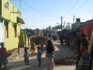 A sunlit village street lined with thatched-roof huts and a yellow-green painted house on the left. Several children stand and walk along the dusty concrete lane, including a girl in a light blue top and long skirt in the centre and a small boy in a blue shirt and shorts. Women in colourful saris move about in the background near a row of hanging clothes, and utility poles with overhead wires run across the sky.