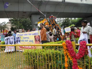 Outdoor street scene beneath a concrete overpass where a crowd gathers along a yellow railing. A large yellow banner held by a man in an orange shirt reads in Kannada, with a portrait of a smiling woman in an orange headscarf printed on it. Other hand-held placards show black-and-white portraits of a woman. A raised decorated float or vehicle is covered with orange and yellow marigold garlands and red flower strings. Women in saris and children stand among the people, and a man in a white shirt holds a white placard at right. Red and orange marigold garlands hang over the railing in the foreground.