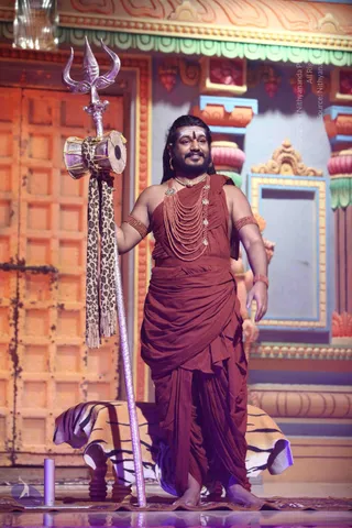A man stands barefoot on a stage in front of an ornate temple-style backdrop with orange carved panels and painted pillars. He wears a rust-red draped garment over one shoulder and a tiered string of coral-coloured beads across his chest with silver pendants. He holds a tall silver trident with a small damaru drum and a leopard-print cloth tied to its shaft. Beside him a striped tiger-skin cloth lies over a low wooden stool. He has long dark hair, a red bindi and a smile. Only one person is visible.