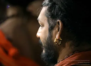 Close side profile photograph of a bearded man with dark hair swept back, wearing a large gold hoop earring with ornate gold clasp detail and a dark rudraksha-style mala around the neck over orange-saffron clothing. Blurred figures in orange robes stand in the background. Dim, warm-toned indoor setting.