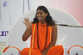 A seated man in saffron-orange robes with gold borders sits on a white cushioned throne in front of a pale backdrop. He wears a long rudraksha mala that hangs to his waist and a small red bindi on his forehead. His long dark hair falls past his shoulders, and his right hand is raised, palm open, as he speaks, with a microphone on a boom arm near his face. Partial white cushions with orange edging appear at the bottom corners. Swamiji (probable): the long swept-back hair, saffron robes with gold borders, and rudraksha mala are consistent with the page's reference, but the image alone does not confirm identity.