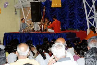 A concert on a raised stage draped with blue pleated fabric. A seated woman in a dark sari plays a tanpura-style string instrument, and a man in an orange kurta sits beside a harmonium-like instrument. A man in grey kurta plays tabla at the left edge. In the foreground, a crowd of seated audience members, mostly with their backs to the camera, including grey-haired and balding men and women with braided hair in white and yellow clothing. A figure in orange clothing sits at the right edge of the stage. Overhead a yellow cloth hangs from the metal truss.