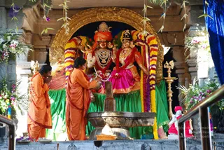 Two priests in saffron-orange robes perform worship before a shrine holding a large deity murti of Shiva and Parvati seated together, adorned with gold jewelry, a crown, and a trident. Garlands of orange, yellow, and purple flowers drape the figures, and a green cloth backdrop hangs below. A dark shivalingam stands on a stone yoni-pitha base in front, with brass oil lamps and flower decorations around the altar. The taller priest at the center pours or offers from a vessel while the other stands with folded hands in a respectful pose.