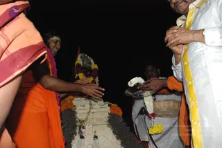 Night-time photograph of a ritual at a white decorated stone structure draped with marigolds, yellow and orange flowers and green foliage. A man in an orange saffron cloth with a pale forehead mark and a pink flower garland reaches his hands over the structure. A man in white shirt with a yellow-bordered dhoti stands at right with hands folded, and a man in white dhoti with an orange cloth holds a silver plate with white and yellow garlands. Dark background.