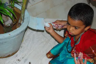A young child in a red and turquoise traditional outfit with a dark bead mala necklace sits on a white cloth and paints a light blue pot with a brush. The child's hand holds a small white dish with red powder, and a palm plant grows in the pot. The setting is a tiled floor. No a person is visible in this scene.