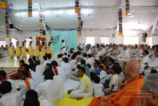 A large white-tented hall with a yellow floor mat and hanging decorative cloth panels. In the left foreground, women in yellow and orange saris with red hair decorations dance in a line near a teal backdrop bearing a green map outline. A man in white clothing plays a hand drum at the centre. Seated audience members in white garments fill the floor, many wearing white garlands, and a row of people in white sit on chairs on the right. A monastic in saffron-orange robes sits in the lower right foreground, and a rudraksha-adorned figure is not clearly visible. The scene appears to be a cultural performance during an event.