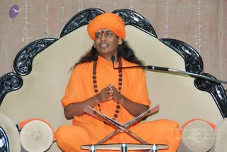 A man seated cross-legged on a cream upholstered throne with an ornate black carved frame sits behind an open book on a wooden X-stand. He wears an orange turban with a red bindi, an orange robe and a long rudraksha mala. His hands are pressed together and a microphone on a stand is beside him. Swamiji (probable): long swept-back hair, orange turban and robe, rudraksha mala. Behind him hang strings of beads on a beige cloth backdrop. A small purple logo appears at upper left.