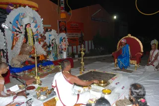 Night-time outdoor ritual before a white and silver decorated deity murti of a goddess with multiple arms holding weapons, seated on a lion, beside other painted deity figures. A figure in a blue robe with a pink-and-white head wrap and beaded necklaces sits on an ornate red and gold throne at right, while a bald priest in white dhoti and sacred thread stands at a square fire pit, pouring from a brass vessel over flower-strewn offerings. Another priest in white sits at left beside a small brass ritual table with lamps and bowls. Brass oil lamps stand on either side of the pit. A red banner with Kannada script hangs behind. Two priests and a seated child are visible; a few other figures appear at the edge.