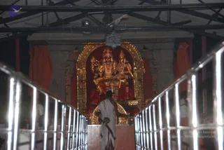 Interior view of a temple shrine seen down a metal-railed walkway. Inside a gilded arched niche stands a large deity murti with a crown and gold ornaments, flanked by a smaller companion figure, draped in red cloth and garlanded with garlands. In the foreground a single priest in white dhoti with a maroon-bordered cloth walks toward the shrine, facing away from the camera. Red curtains hang at the sides, and a corrugated roof with lamps and wiring overhead. Faint figures appear at the edges of the frame.