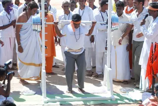 Outdoors on bare earth, a man in a white t-shirt, grey trousers, a leather belt and an orange headband squats beneath a loaded barbell with weight plates, gripping the bar at shoulder height on a white rack. Around him, men in white dhotis and white shirts with rudraksha-style neck malas, one in orange robes, and a man in a white shirt holding a microphone watch closely. A man in a white dhoti with a gold border stands at left, and orange flags are visible at right. A green mat lies beneath the rack, and a photographer with a camera is at lower left.