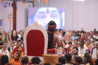 A seated figure with long braided dark hair tied with a rust-orange headwrap sits on an ornate silver-and-red throne seen from behind, facing a large projection screen showing a close-up face with a red tilak on the forehead. A silver trident stands to the left of the throne. Rows of seated devotees fill the hall, many in orange, red and white clothing, some wearing rudraksha malas. A small table with a microphone sits in front of the throne, and a man in white with a mala sits near the foreground at right.