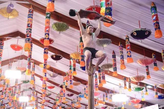A shirtless man with a black headband and dark shorts balances on top of a tall wooden pole while holding a second person's body horizontally above him in one raised arm. Beneath the ceiling, hung with many colourful decorated cylindrical and umbrella-shaped ornaments in orange, red, blue, purple and green, two bright white lamps glow. The setting appears to be a large festive tent or pandal. Only the man on the pole and the partially visible person being held are physically present.