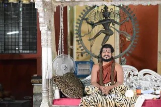 A bearded man with dark shoulder-length hair sits cross-legged on an ornate silver-framed couch with a red cushion, smiling. He wears a rust-orange wrap over bare shoulders, a long rudraksha-style mala necklace, and tiger-striped trousers, with a red tilak on his forehead. Behind him stands a bronze Nataraja statue of dancing Shiva within a flame arch, beside a silver-pillared canopy with hanging chains and string lights. A leopard-print bolster rests beside him, and a fan and ritual items sit nearby. The setting appears to be a temple-style hall with red walls.