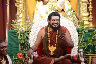 A smiling man with long dark hair and a full beard sits cross-legged on an ornate white and red throne, looking down at a smartphone held in his right hand. He wears a rust-orange robe, a thick rudraksha mala and a large gold ceremonial pendant necklace, with a white tripundra marking on his forehead. His left hand rests near a silver vessel on a tray. Behind him, gold-yellow fabric and gilded brass lamps frame a green cloth and potted plants. At the left edge, a smiling man in white ritual clothes with a gold-bordered shawl sits beside a tall gold staff.