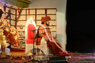 A seated man on an ornate silver-and-red throne speaks into a microphone on a stage draped with a colourful striped woven cloth. He wears a rust-orange turban, orange-brown robes, a pink-and-white shawl, gold necklaces and a rudraksha-style bead mala, with a long braid of hair falling over his shoulder. A lit oil-lamp stand sits beside him, and a wooden lattice behind is strung with red flower garlands and small lights. To the left, a life-size statue of a seated figure in a black crown and orange-brown dhoti wears layered flower garlands and holds a staff. The scene looks like a seated discourse set.