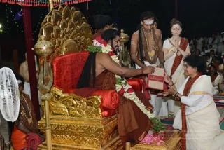 A seated figure on an ornate gold throne with a red velvet cushion and a golden peacock-feather back is garlanded with white and red flower garlands and pink petals at the feet. He wears a rust-brown robe and has long dreadlocked hair. A woman in a white and cream sari with a red-bordered shawl kneels at right, offering a small red-and-gold packet, while a woman in white with a red shawl stands behind her with palms pressed together. A man in white dhoti, bare-chested with white forehead markings, a brown shawl and gold necklace stands behind the throne. Golden trident-topped staffs, a white fan at left and a seated attendant in orange garments frame the scene. Several people in white are seated in rows in the background under strings of lights and a red canopy.