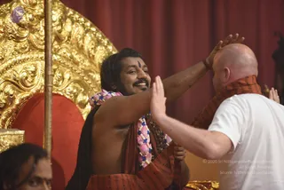 A seated man with long dark hair and a beard, bare-chested with a rust-orange shawl draped over his shoulders and a pink and blue lotus garland, smiles while raising his hands to bless a bald man in a white t-shirt who bows before him. A gold ornate throne with a red cushion stands behind him against a red curtain, and a gold staff rises at left with a partial figure in the foreground. Swamiji (probable): the seated man has long swept-back hair, a bindi-style tilak, rudraksha mala beads and ceremonial lotus garland, seated on a golden throne receiving devotee obeisance.