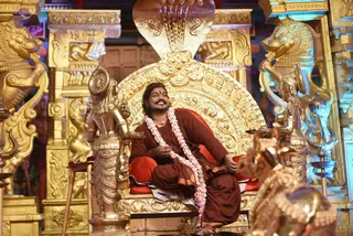 A man with long dark hair sits on an ornate gold throne with a cobra-hood canopy and a red cushion. He wears a rust-brown robe and a long white and pink flower garland. Golden lion and deity figures flank the throne, with lamps and a small table with flowers at left. The page indicates a person is present, but the appearance cues are limited to hair and garland, so identity is uncertain.