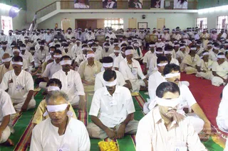 A large hall filled with many men seated cross-legged on green and striped mats, most wearing white shirts and white dhotis or mundus and white cloth blindfolds tied over their eyes. Some wear lanyard ID badges. Several seated in the foreground have hands resting on their laps, and a few wear saffron-yellow or cream shawls. Red carpet covers the right side of the floor. Above, on an upper balcony, framed portraits hang along the railing; the portraits are printed images, not physical people.