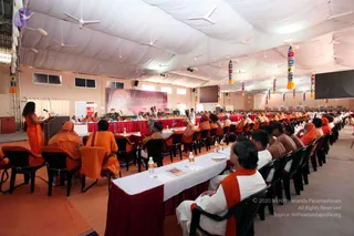 A wide view of a large white-canopied hall during a seated assembly. Rows of attendees in saffron and orange robes sit at long tables draped in maroon cloth, with white table runners and water bottles. A woman in an orange sari stands at a wooden podium on the left with a microphone. Dozens of people in saffron and white clothing fill the rows, and a banner reading Anandotsavam hangs on the back wall beside a portrait. Colourful tasselled hangings decorate the ceiling, and white ceiling fans are visible.