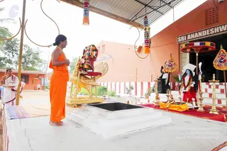 Outdoor temple courtyard with an orange-painted wall and a sign reading Sri Anandeshwari. A man with long hair tied up, wearing an orange saffron robe, flower garland and bare feet, stands with hands folded in front of a golden peacock-shaped throne holding a flower-draped deity murti. Beside the murti stands a blue-skinned goddess figure with red garments and a crown, and a white-clad priest in the background holds a tray. Gold oil lamps, a kalasha-like decorations, colourful hanging tassels and a square fire pit or homa altar sit on the platform. Ritual worship scene in an ashram setting.