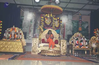 A seated figure in saffron-orange robes sits with palms pressed together on an ornate gold throne with red cushioning, before a stage set with gold-framed thrones, garlanded deity murtis and marigold garlands. A central golden peetha-style seat is backed by a large decorated umbrella-canopy and floral garlands framing two deity murtis. Flanking thrones hold garlanded figures and a golden lion-like sculpture appears beside the central seat. Floral and orange draped cloth covers the platform, with a large backdrop of stone temple pillars and a painted forest scene. A rudraksha-style mala or ceremonial adornment is not clearly visible. The seated figure's appearance shows long dark hair and orange robes, with a white-shawled figure in the central position.