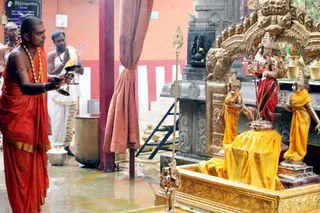 A photograph inside a temple shrine where a priest in saffron-orange robes with a rudraksha-style bead necklace and bangle holds a lit oil lamp aloft toward golden deity murtis. Behind him a second man in white dhoti and shawl stands with hands joined in prayer. The golden murtis, dressed in yellow and red garments with a spear-like staff, sit beneath an ornate gold arch against a granite wall, with a small dark murti and marigold offerings on the upper shelf and brass kalashas nearby. Two other men stand at the left edge. This appears to be an aarti during a puja.