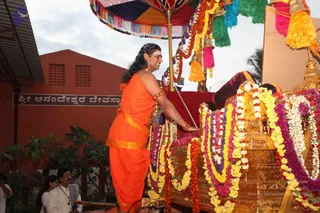 A photograph of an outdoor festival procession at a temple-style building with a red-brick wall and a Kannada signboard reading Sri Anandeshwara Devasthana. A man in a saffron-orange dhoti and shawl, with long swept-back hair, a white tilak on the forehead and multiple beaded and gold necklaces, stands on a golden ornate ratha/chariot, one hand on its rail. The chariot is heavily garlanded with orange, yellow, purple and white flower garlands and draped with red velvet, beneath a multicoloured canopy with gold tassels and a kalasha-style finial. Two or three onlookers in white stand at the lower left and a woman in a sari is partly visible behind them.