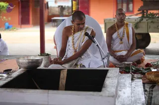 A homa or yagna is being performed at an open-air ritual area with a square stone fire pit in the foreground. Two men in plain white ritual cloth with yellow-gold borders sit cross-legged on a raised platform. The man in front has a shaved head, a gold-coloured bead necklace, and his hands folded over a sheet of paper. A microphone stands beside him. The man behind him, also shaved-headed and wearing glasses and a gold necklace, sits with hands joined. Brass bowls, a leafy plant, a brass lamp, fruit and a garland are arranged on the platform. An orange building and a cart are visible behind.