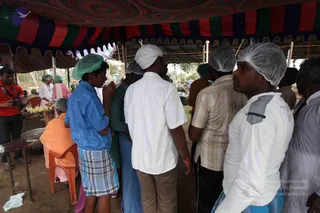 A photograph of an outdoor food-service area under a colourful striped tent canopy with red, green, blue and maroon bands. In the foreground, several men queue in a line wearing white and green disposable hairnets: one in a blue shirt and checked lungi, one in a white shirt and white head cloth, one in a beige checked shirt, and one in a white shirt with a checked panel. Behind them, a woman in a white hairnet and a woman in orange-pink sari stand near a table with plates and food. A seated person in orange clothing sits on a red plastic chair at left, and a photographer in a red shirt with a camera stands at the left edge.