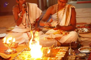 A homa fire ritual on the floor. A young priest in white dhoti and shawl with a gold-toned trim speaks into a microphone on a stand at left. A seated priest in white dhoti and shawl with gold border, wearing glasses and a gold bead necklace, holds a brass plate of offerings over a blazing fire pit of wooden logs. Brass oil lamps, a bell and silver vessels sit nearby on the floor.
