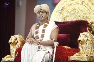 A man seated on an ornate golden throne with carved lion-head armrests and a red velvet cushioned seat with a zebra-stripe pattern. He wears a cream turban with a jewelled crest, a red bindi, a cream dhoti and shawl, and layered rudraksha beads over the chest, with a red-and-gold armband. He wears a headset microphone and speaks while resting his hands on his knee, with a white staff in his right hand. A golden canopy with ornate metalwork rises behind him. The a person label is unsure because the appearance cues are not strong enough to confirm a candidate.