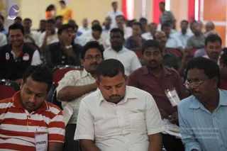A photograph of an indoor satsang hall with rows of seated men in red chairs, many with eyes closed in meditation. In the foreground, a man in a red-and-white striped polo with a PRESS badge sits beside a man in a white shirt with a pen-holder badge and a man in a maroon shirt and a man in a light blue shirt with glasses. Behind them, many more attendees fill the hall, some smiling, one in a navy vest with a number 8 on it. Pale orange walls and red drapes frame the room. No teacher is visible in this view.