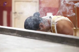 A photograph of a man in a white dhoti with a gold-bordered cloth bending low over a stone ledge, his head and shoulders partly obscured by rising smoke from incense or a sacrificial fire. He wears a red-brown rudraksha mala around his neck and a white upper cloth. His dark hair is tied in a knot at the top of his head. A brass vessel holding incense sticks sits at the right edge, and a red wall and pale doorway form the blurred background. The man appears to be in a devotional posture, such as a prostration or offering. The scene is a single person in a temple or ashram setting.