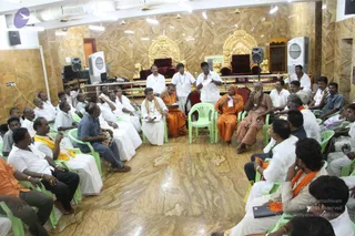 A seated group of roughly two dozen men gathers in a circle in a hall with gold-veneered marble walls and two ornate gold thrones with red cushions at the back. Most wear white dhotis and shirts, while several in saffron/orange robes sit near the centre, including one holding a microphone and one with a long grey beard and rudraksha mala in reddish-brown robes. Green plastic chairs are arranged in rows, and a standing man in a white shirt holds papers and a microphone. Floor-standing speakers and wall air coolers are visible.