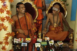 Two seated men in saffron-orange robes sit on ornate gold-framed thrones with dark maroon cushions, microphones from multiple television and news channels pointed at them. The man on the left, smiling broadly, wears a saffron wrap over his shoulder, rudraksha malas, a silver necklace and a red headband, with a red bindi on his forehead. The man on the right, bald and bare-chested, wears a white sacred ash band on his forehead, a red headband, rudraksha malas, a gold chest ornament and gold bangles, and gestures with his hands. Behind them stands another robed figure in orange, and autumn-leaf garland decorations frame the scene against a blue-and-white wall.