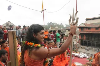 A woman with long dark hair, wearing a saffron-orange garment and a marigold garland with a rudraksha bracelet, raises a silver trident in her right hand at an outdoor temple courtyard. Behind her, a seated group in orange robes and a seated devotee sit on a red cloth with metal plates and offerings. A crowd of onlookers stands in the background, and a cameraman in a green shirt films beside a tripod. Red brick temple buildings, a yellow flag on a pole, and distant spires are visible. The scene suggests a puja at a temple. No printed captions or readable text appear.