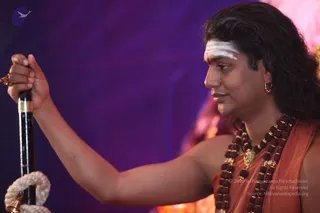 Photograph in profile of a man holding a long black wooden staff with a white rope coiled at its base and a small brass bell hanging near the bottom. He wears an orange-rust draped garment, multiple dark rudraksha mala strands, and a gold pendant necklace with a small gold ornament and a gold earring. A white horizontal tilak with a red dot marks his forehead. The background is a deep violet backdrop with warm pink light behind him. Swamiji (probable): long swept-back hair, rudraksha malas and ceremonial gold necklace visible, consistent with the page's identification of Swamiji at this satsang.