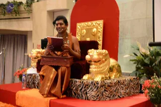 A photograph of a seated figure on a raised red-carpeted stage, reading from an open red book held in both hands. The person wears an orange-saffron dhoti with a gold-bordered cloth draped over the shoulder, bare-chested, with a gold ornate necklace, a gold armlet, a red bindi, and a small headset microphone. Behind is an ornate gold throne with a deep red velvet back and gold lion-form armrests set on a tiger-striped plinth. Orange and red flowers and green foliage decorate the platform sides, and a rectangular wooden stand rests in front. Swamiji (probable) is the seated figure: long swept-back hair, ornate gold necklace, saffron clothing and a crown-like throne setting, presenting a discourse.