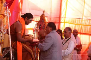 A seated figure in orange and ochre robes with a long swept-back hair bun and white forehead marking receives an offering from a man in a light grey jacket. The seated figure wears an orange shawl, gold armband and bead necklaces, and sits before a silver trident and a gold-framed throne. Behind them stands an attendant in a red and gold cloth, while to the right a man in a white kurta with a lanyard, a woman and a bald man in a white shawl watch. Orange and yellow draped fabric forms a tent-like backdrop. Swamiji (probable): the seated figure has long swept-back hair, orange robes and ceremonial gold adornments on a throne.
