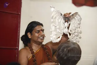 A man in a saffron-orange shawl wrapped over his bare shoulder, wearing a large rudraksha mala with a gold-edged dark bead chain, speaks while holding up an ornate silver ceremonial object shaped like a crescent with scrolled arms and round finials. A small red bindi marks his forehead and his long dark hair is swept back. A second person's head is visible at the lower right, and a red wooden door stands at the left against a cream wall. The scene reads as a teaching moment with the silver object used as a visual aid.