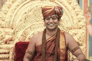 A smiling man sits on an ornate gold throne with a red tiger-striped cushion. He wears a rust-orange turban with a jewelled ornament, orange-saffron robes draped over one shoulder, long rudraksha mala necklaces, bead bracelets on his upper arms, and a red bindi. A small microphone is clipped near his face. Behind him is a carved gold arched backdrop. Swamiji (probable): long turban, saffron robes, rudraksha malas and ceremonial jewellery, seated as a teacher on a throne.