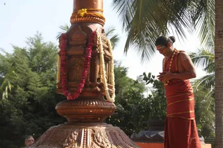 A photograph outdoors under palm trees and greenery shows a bare-chested man in a rust-orange dhoti with a gold-edged cloth draped over his shoulder and a rudraksha mala around his neck, standing with palms pressed together in namaskar and head bowed. Beside him stands a tall copper-coloured ceremonial pillar or kalasha-style lamp stand with carved panels and a base, draped with red and white flower garlands and a yellow cord, with scattered petals on the ground. A small figure in the lower left edge is partially visible.