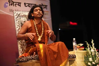A photograph of a man seated on an ornate silver-carved throne-style chair during a talk on a stage, gesturing with both hands while speaking into a headset microphone. He wears a saffron-orange robe with a gold-bordered cloth, rudraksha mala beads and a heavy gold necklace, with a red bindi on his forehead and gold bangles. A leopard-print cushion rests on the chair. Beside him stands a small table with a water bottle, white lilies and red roses. Behind him is a red-and-cream banner with a purple logo and the printed words "Nithya Dhyan Yog" and "life bliss". The stage is dark with a red light in the background. Swamiji (probable) is seated centrally, identified by long swept-back hair, rudraksha malas, ceremonial gold necklace and saffron robe.
